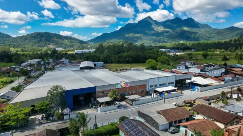 Vista aérea de uma fábrica em área rural, com galpões industriais e pátio ao lado de casas, cercada por vegetação verde e montanhas ao fundo sob céu parcialmente nublado.